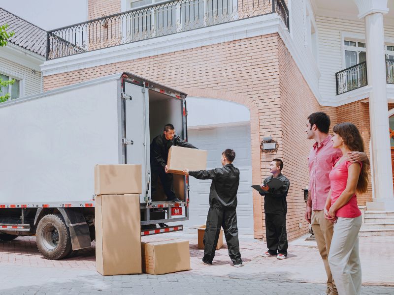 A moving company is loading boxes into a truck while a couple watches in front of their house.