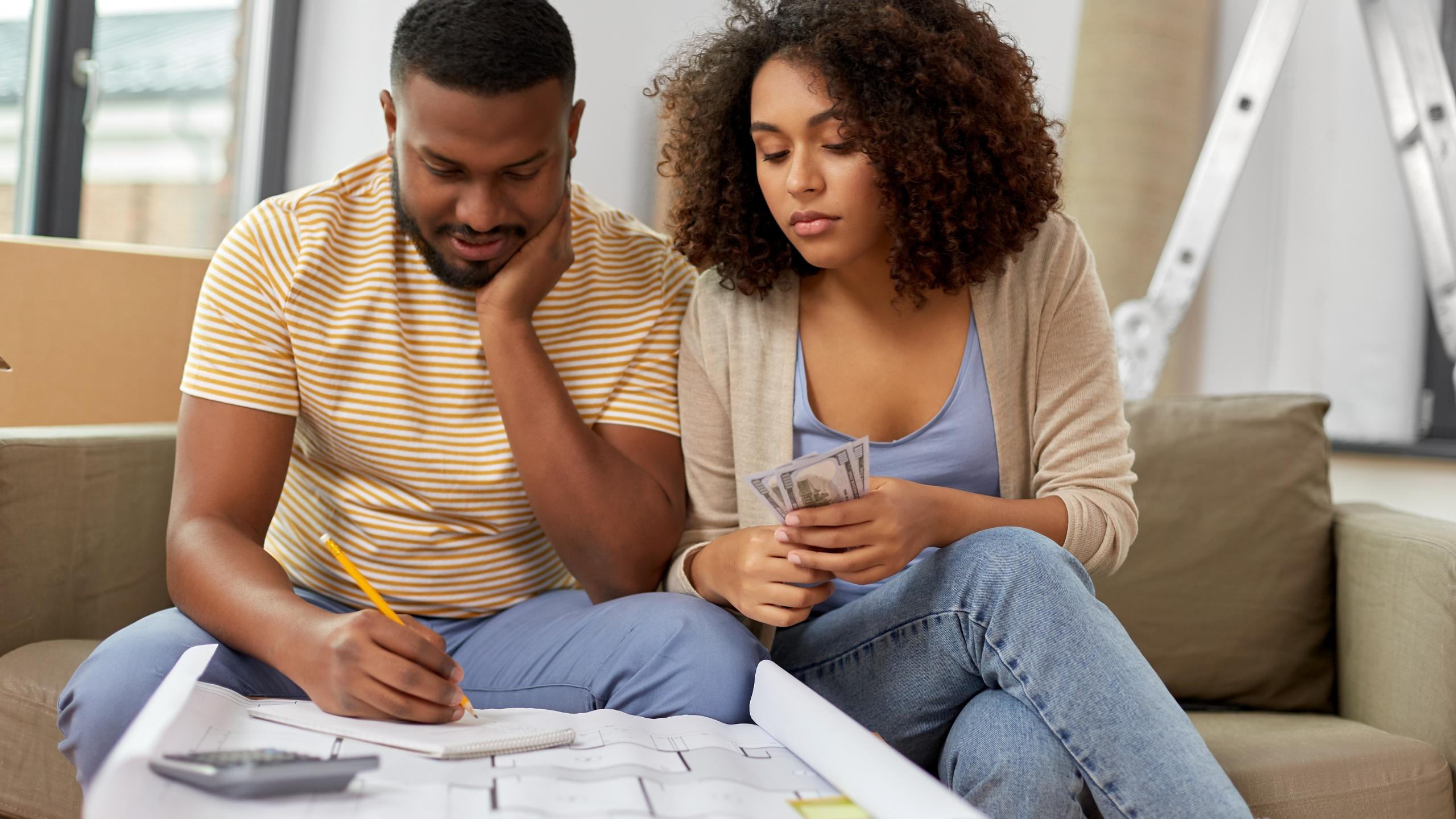 A couple sitting on a couch, looking at blueprints and money while planning their move.